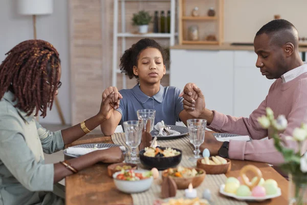 Portrait of modern African-American family saying grace at dinner table on Easter and holding hands, copy space