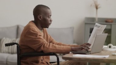 Orta boylu, yakışıklı, özürlü bir Afrikalı iş adamının oturma odasında, elinde bir belge diğeriyle birlikte dizüstü bilgisayarda klavyeyle otururken görüntüsü.