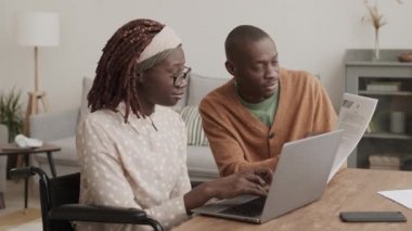 Genç Afrikalı bir adam elinde belgelerle masada otururken Afrikalı engelli bir kadının tekerlekli sandalyede oturup dizüstü bilgisayarda klavyeyle yazarken orta boy bir fotoğrafı.