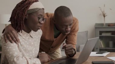 Orta boy Afro-Amerikalı bir adam, çekici kız arkadaşının yanında dizüstü bilgisayarın önünde oturuyor, tek eliyle ona sarılıyor ve dizüstü bilgisayara bakıyor ve konuşuyor.