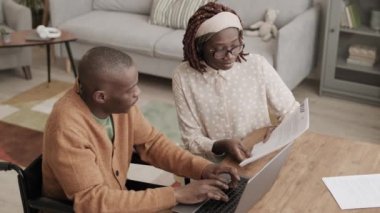Genç Afrikalı kadın elinde belgelerle masada otururken Afrikalı engelli bir adamın tekerlekli sandalyede oturup dizüstü bilgisayarda klavyeyle yazı yazdığını yüksek açılı görüyor.