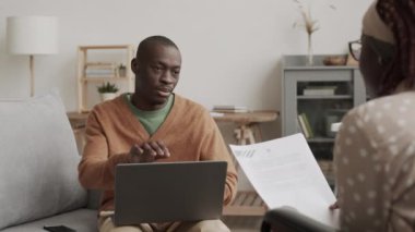 Genç Afrikalı bir adamın oturma odasında kanepede oturup dizüstü bilgisayarla işini bitirirken önünde elinde belgelerle oturan genç Afrikalı bir kadınla sohbet ederken orta boy fotoğrafı.