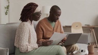 Genç Afrikalı bir kadınla bir adamın kanepede oturup evrak işleriyle uğraşırken orta boy fotoğrafı. Dizüstü bilgisayarda klavyede yazan adam