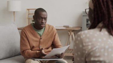 Elinde belgelerle kanepede oturan Afro-Amerikalı bir adam ve önünde oturan tanınmayan Afrikalı bir kadınla öfkeyle konuşuyor.