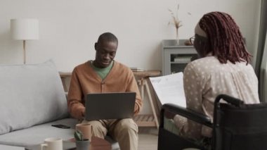 Genç Afrikalı engelli bir kadının tekerlekli sandalyede elinde belgelerle oturup Afrikalı bir adama kanepede oturup dizüstü bilgisayarda klavyeyle yazı yazarken orta boy bir fotoğrafı.