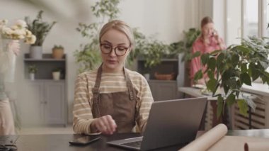 Sarışın, beyaz tenli, gözlüklü, çiçekçi masasında oturan, dizüstü bilgisayar ve akıllı telefon kullanan, arka plan üzerinde çalışan,