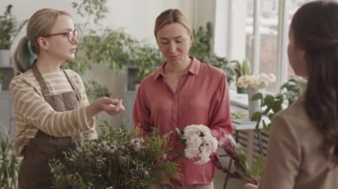 Kafkasyalı iki kadın çiçekçi dükkanın önünde dikilip konuşuyor ve tanınmayan müşteriye bitkiler sunuyorlar.