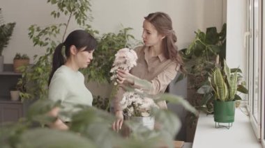 Orta uzunlukta beyaz bir bayan yönetici yeni Asyalı çalışanı fabrikada eğitiyor, çiçeklerini gösteriyor, işini anlatıyor.