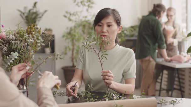 花の店で机に座っている若いアジアの女性花屋のウエストアップ セクターで装飾植物の枝をトリミングし 認識できない同僚の意見を求める — ストック動画