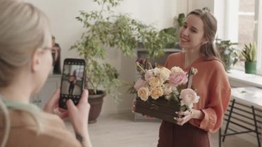 Sarışın, beyaz bir kadının, elinde çiçek aranjmanıyla ahşap sepet tutarak genç bir kadının fotoğrafını çekmesi, çiçekçinin önünde durması, sonra da resmini göstermesi.