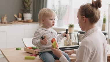 Annenin evde çalışıp dizüstü bilgisayardan online konferans izlemesi masasında oturan sarı saçlı bebekle oynaması.