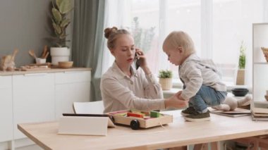 Evde çalışan, cep telefonuyla konuşan, tablet bilgisayar kullanan, masa başında duran şirin bir çocuğa bakıcılık yapan beyaz bir genç.
