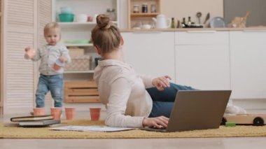 Genç, meşgul, beyaz bir annenin dizüstü bilgisayar kullanarak yerde yattığı, sarı saçlı bebeğinin arka planda fincanlar taşıdığı bir sahne.