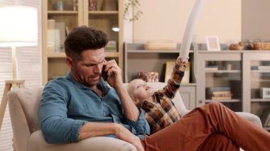 Orta boylu, mavi gözlü, beyaz bir adam oturma odasında oturuyor, akıllı telefonla konuşuyor, iki yaşındaki oğlu babasının yanında yumuşak kılıç oyuncağıyla oynuyor.