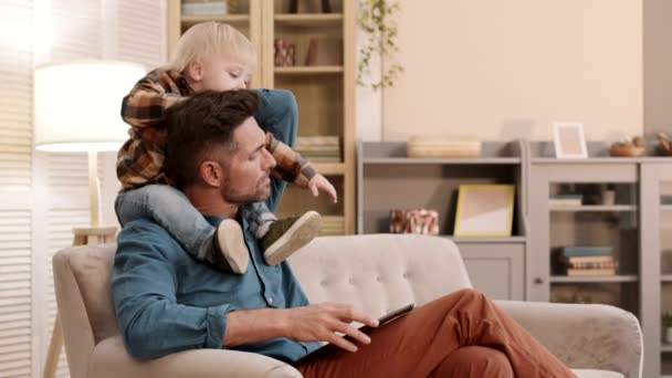 中等个子的白人男子坐在家里的沙发上 用平板电脑 两岁的孩子肩膀上坐着 与父亲的头玩耍 — 图库视频影像