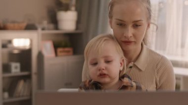 Evdeki genç ve güzel kadının orta boy yakın çekimi, kollarında tatlı gözyaşı lekeli bebeğiyle, onunla konuşuyor.