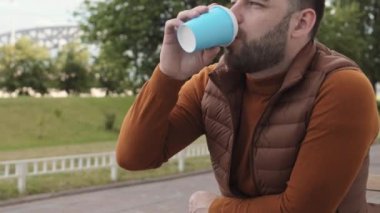 Kahverengi saçlı, kahverengi kazak ve yelek giyen, açık kafeterya masasında kitap, akıllı telefon ve kulaklıkla oturan, parkın yanındaki mavi kağıt bardaktan kahve içen genç bir beyaz adam.
