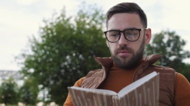 Gözlüklü, kitap okuyan, kağıt bardaktan kahve içen ve kameraya bakıp gülümseyen neşeli, beyaz bir adamın düşük açılı görüntüsü.