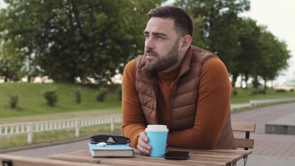 身着棕色毛衣和背心 头戴棕色头发的年轻白种人站在公园附近的户外咖啡馆桌旁 用蓝色纸杯喝咖啡 — 图库视频影像