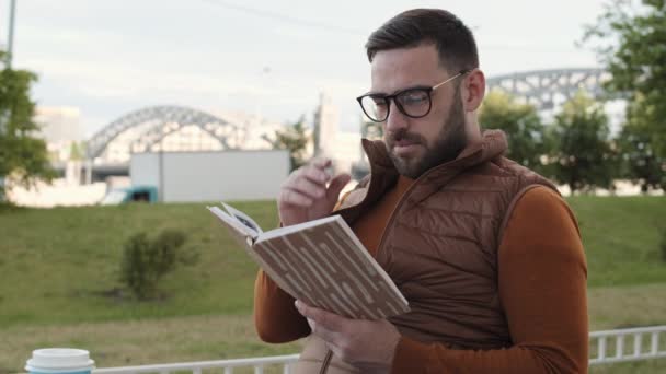 一个年轻英俊的白种人站在公园附近的一座桥边 一边看书 一边举杯 一边笑着 站起身来 扬起了一个弧形的弧线 — 图库视频影像