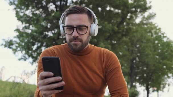 一个快乐的棕色头发的白种人 头戴白色无线耳机 听音乐 走起路来 手里拿着智能手机 坐在公园里 — 图库视频影像