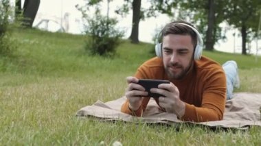 Kahverengi süveter giyen, kablosuz kulaklık takan, yeşil çimlerin üzerinde battaniyenin üzerine uzanmış, akıllı telefondan oyunlar oynayan, gülümseyen ve kazanan beyaz bir adam.