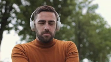 Kulaklık takan, müzik dinleyen, hareket eden, gülümseyen ve parkta kameraya bakan genç, sakallı, beyaz bir adam.