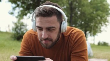 Kahverengi süveter giyen, kablosuz kulaklık takan, midenin üzerine uzanmış, parkta oyun oynayan, akıllı telefondan gülümseyen ve kazanan orta boy beyaz adam.
