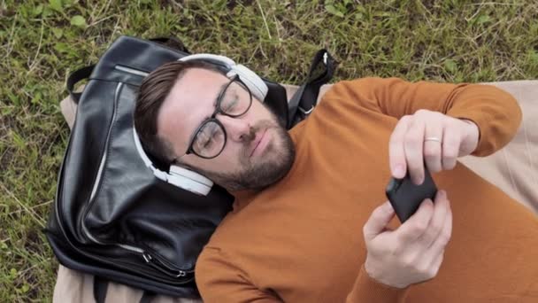 안경을 스웨터를 헤드폰을 스마트폰을 사용하여 잔디밭에 가방에 머리를 남자의 — 비디오