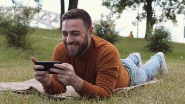 许多快乐的白人年轻人穿着棕色的毛衣 躺在公园草坪上的毯子上 用智能手机 然后微笑着接听电话 然后转身离去 — 图库视频影像
