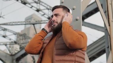 Düşük açılı, neşeli, sakallı, beyaz bir adam. Açık havada, gri metal köprü zemininin önünde. Gülümsüyor, kulaklıkla müzik dinliyor.