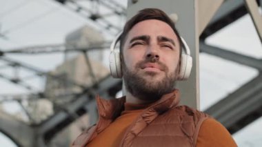 Düşük açılı göğüs yukarı, neşeli sakallı beyaz adam açık havada duruyor, bulanık gri metal köprü zemininde, gülümsüyor, kulaklıkla müzik dinliyor, hareket ediyor.