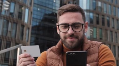 Modern binaların ön planında oturan, akıllı telefondan çevrimiçi ödeme için kredi kartı numarasını dolduran, sonra kameraya bakıp gülümseyen genç beyaz adamın düşük açılı pozu.
