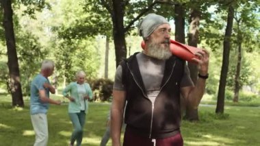 Orta boylu, beyaz sakallı, spor giyimli, şapkalı, yoga minderli orta yaşlı bir adam. İnsanlar arka planda ısınırken kameraya bakıyor.