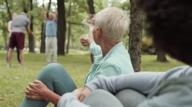 Çekici, yaşlı, kısa saçlı, parkta çimenlikte oturan ve önünde oturan tanınmayan bir kadınla konuşan, arka planda egzersiz yapan, sporcu bir kadın.
