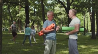 Eğitimden ve sohbetten sonra parkta yoga minderleri tutan iki beyaz erkek yoganın geniş açılı görüntüsü.