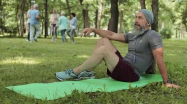 Yaşlı sakallı, beyaz tenli bir sporcunun parkta çimlerin üzerinde oturması ve yoğun eğitimden sonra bir grup son sınıf öğrencisinin arka planda egzersiz yapması.