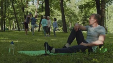 Orta yaşlı yakışıklı beyaz tenli sporcunun parkta çimlerin üzerinde oturup, yoğun antrenmandan sonra dinlenmesi ve arka planda egzersiz yapan bir grup yaşlıyı izlemesi.