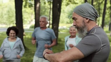 Bir grup yaşlı sporcu parkta egzersiz yaparken orta yaşlı erkek eğitmenin ıslık çalıp saatine baktığı bir fotoğraf.