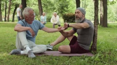 Geniş açıda, çimenlikteki lastik paspasın üzerinde oturan ve antrenmandan sonra yakınlarda oturan yaşlı bir sporcunun nabzını ölçen yaşlı bir erkek koç.