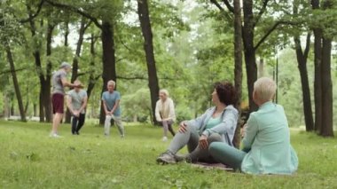 Geniş açıda, spor kıyafetleri giymiş iki güzel kadının parkta çimlerde oturup sohbet ederken bir grup insanın arka planda egzersiz yaptığı görüntüler.