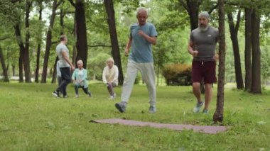 İki yaşlı sporcunun parkta lastik paspasa doğru koşarken, insanlar arka planda antrenman yaparken oturup konuşmaya başladıkları geniş bir çekim.