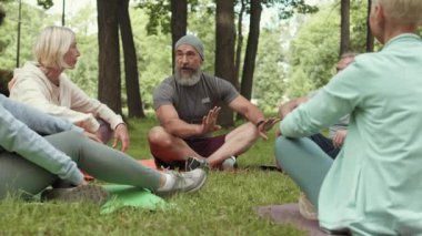 Yaşlı sakallı erkek antrenörün parkta çimlerin üzerinde oturup bir grup üst düzey sporcuyla konuşurken çekilmiş bir fotoğrafı.