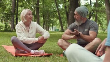 Bir grup yaşlı sporcunun parktaki çimlerde oturup antrenmandan önce konuştuğu arka plan görüntüsü. Sakallı erkek antrenör yakınlarda oturan güzel sarışın sporcuyla sohbet ediyor.