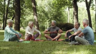 Yaşlı, sakallı bir erkek antrenörün parkta çimenlikte oturup yakınlarda oturan ve onları dinleyen bir grup yaşlı sporcuyla iletişim kurması.