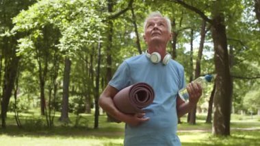 Orta yaşlı beyaz sporcunun spor kıyafetleri ve kulaklıklar takıp ormanda elinde yoga minderiyle durup kameraya bakarken çekilmiş bir fotoğrafı.