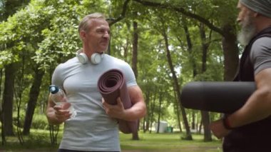 Orta yaşlı, orta yaşlı, beyaz bir sporcunun parkta duran kulaklık taktığı ve yaşlı sakallı bir sporcuyu selamladığı bir fotoğraf. Kameraya bakıyorlar.