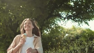 Orta boylu, neşeli, beyaz bir kadın. Yaz parkında zıplıyor, dondurma tutuyor, gülümsüyor ve kameraya bakıyor.