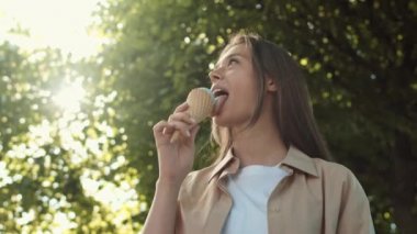 Güneşli yaz parkında duran, dondurma yalayan, gülümseyen ve kameraya bakan genç, mutlu, beyaz bir kadının arka plan pozu.