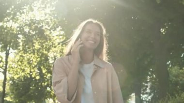 Neşeli, beyaz bir kadının cep telefonuyla konuşurken, gülümserken, güneşli yaz parkında yürürken arka planda çekilmiş görüntüleri.